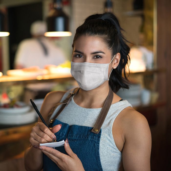 Portrait of a beautiful waitress working at a restaurant wearing a facemask while holding a notepad and looking at the camera â pandemic lifestyle concepts