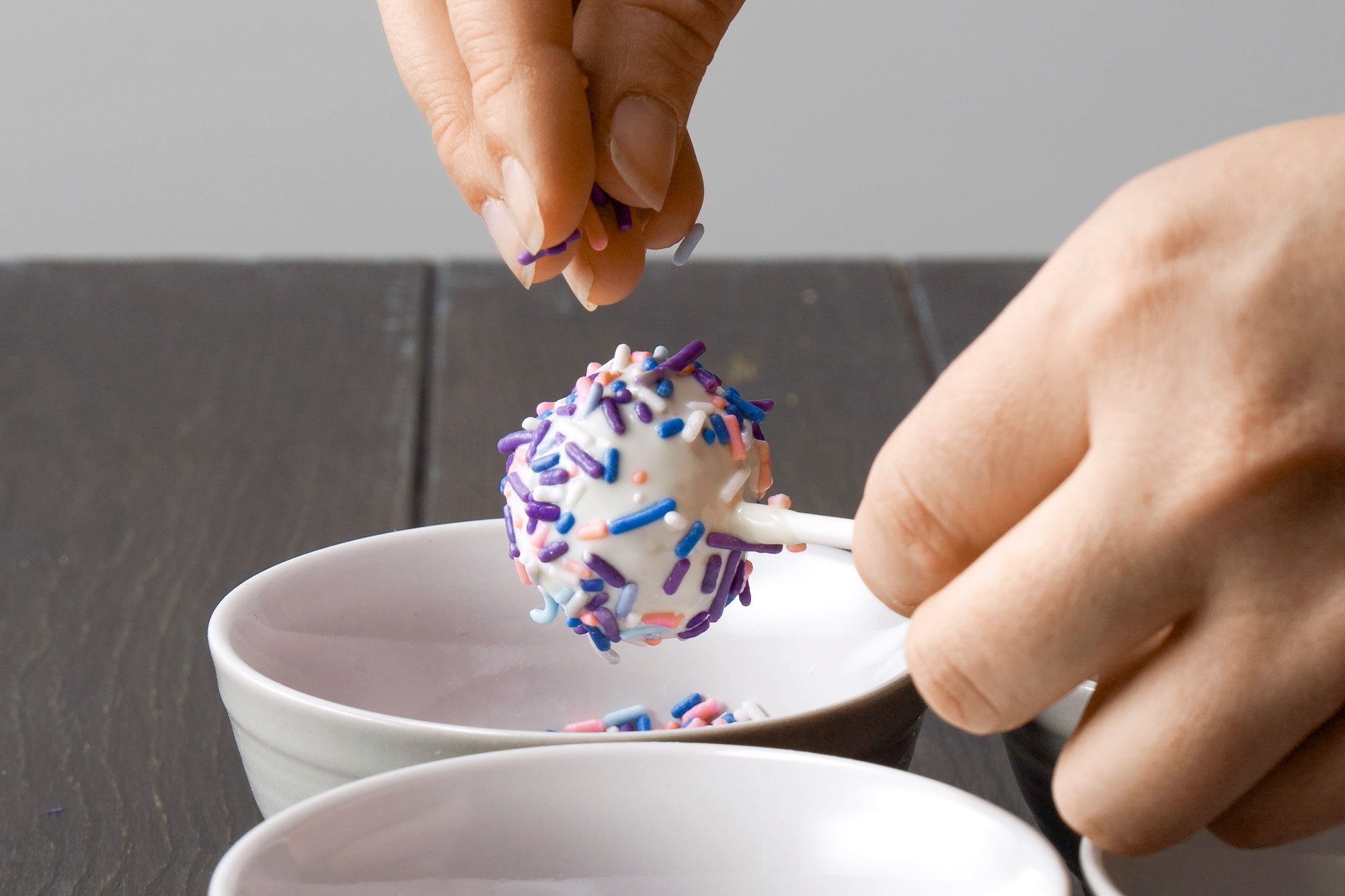 horizontal shot of a hand sprinkling colorful sprinkles onto a cake pop; in the foreground, there are two small white bowls placed on a wooden surface