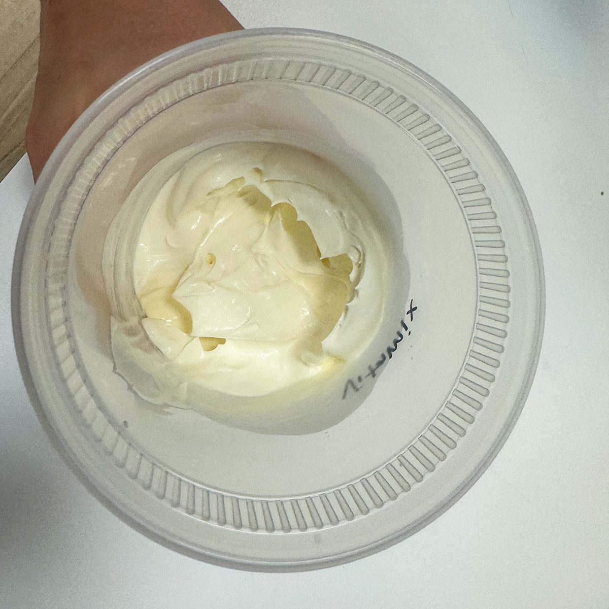 A small plastic container filled with creamy white yogurt sits on a table. The lid is partially visible, and the word "Thickness" is written on the side.