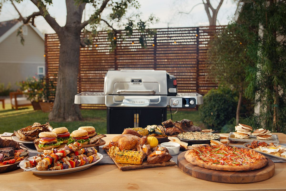 A table outdoors laden with grilled food, including burgers, pizza, corn, skewers, and assorted side dishes. A large grill is in the background, set in a backyard with trees and a wooden fence.