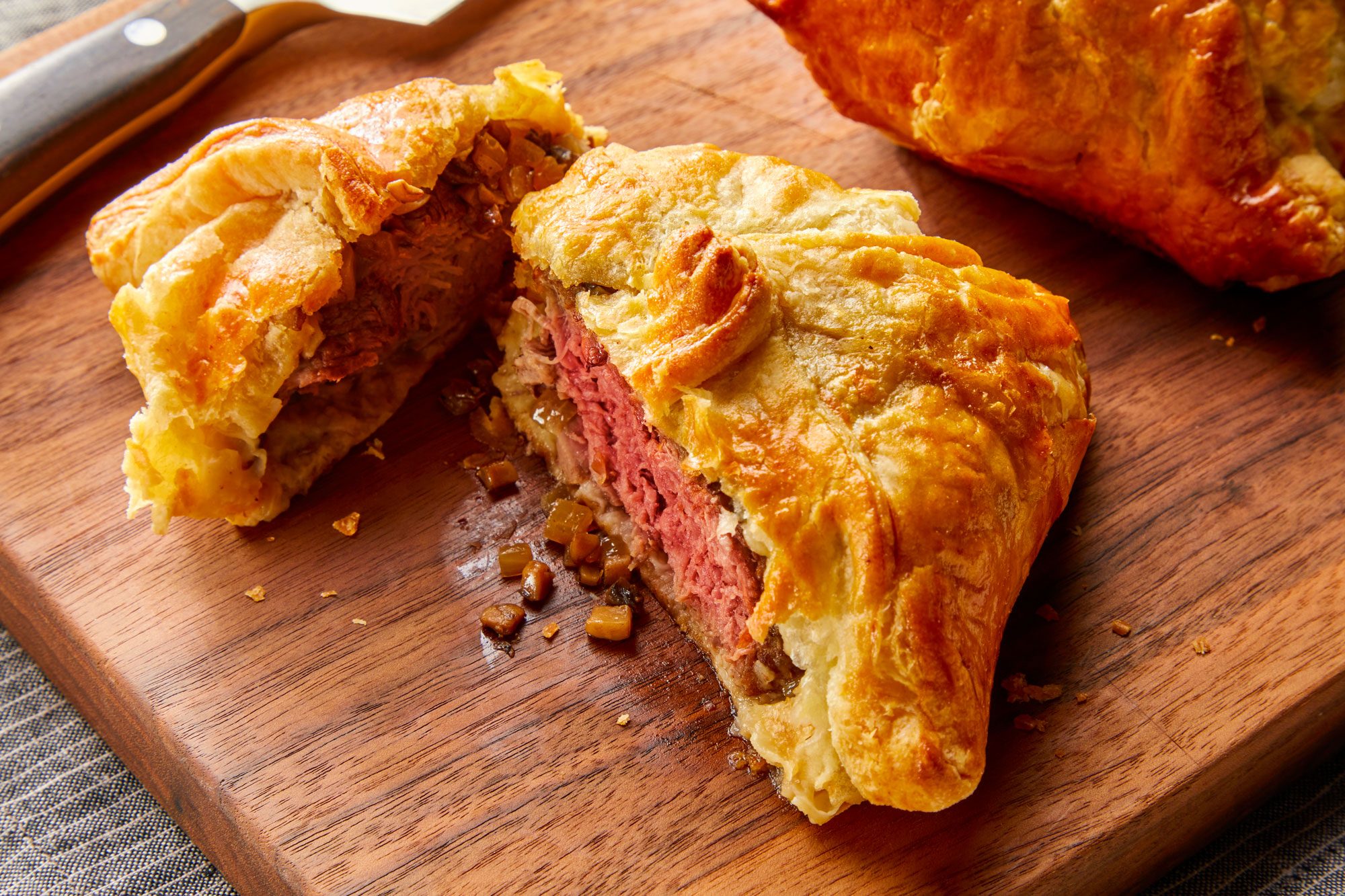 3/4th shot of Beef Wellington resting on a wooden cutting board along with a knife