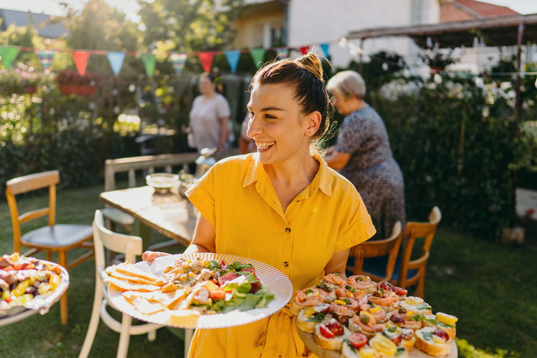 Happy guest bringing two food dishes to an outdoors summer potluck party in a backyard.