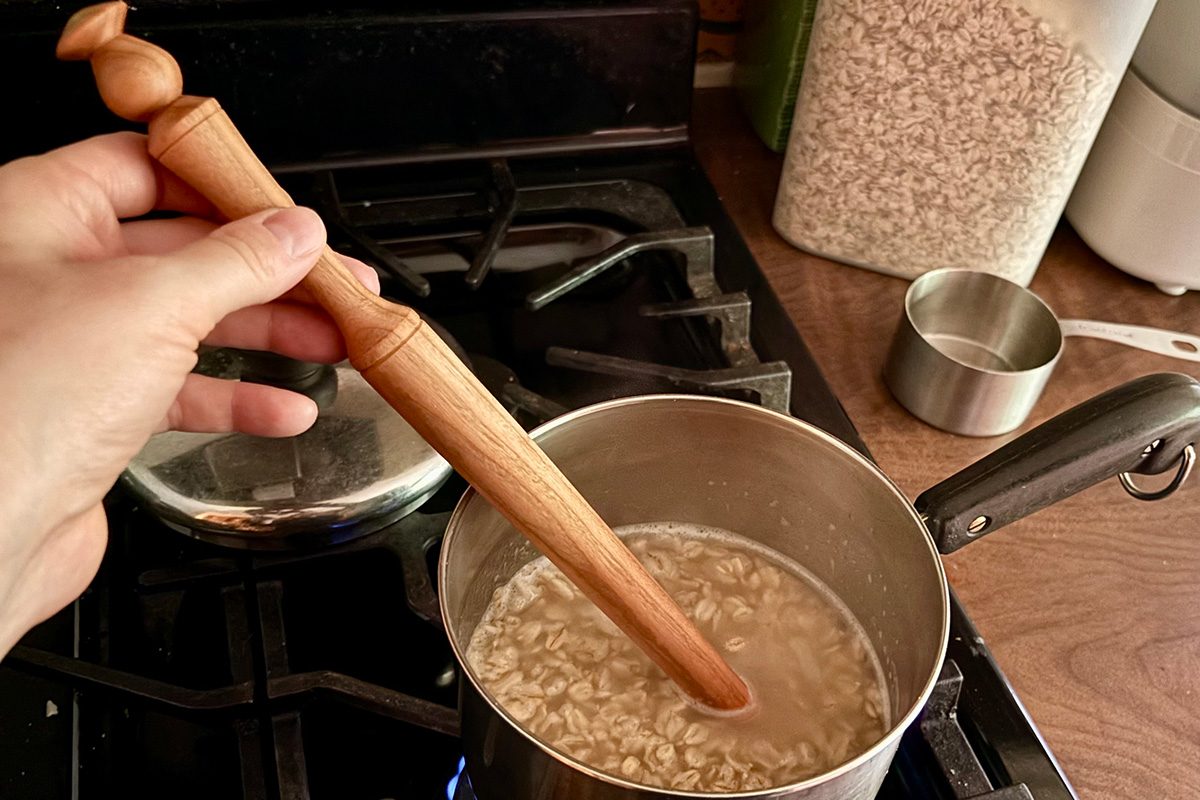 A hand stirs oatmeal cooking in a pot on a stove with a wooden spoon; a metal measuring cup and a container of oats are on the counter nearby.