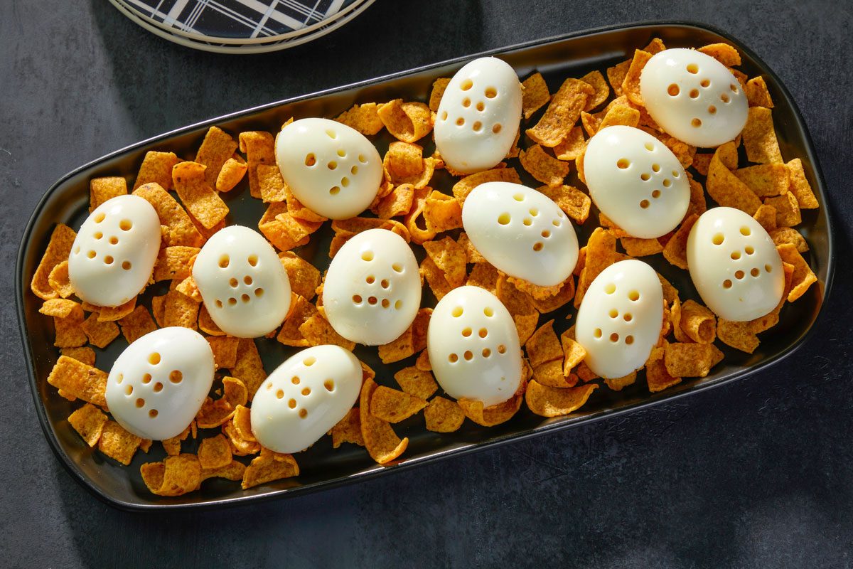 Overhead shot of Skull Deviled Eggs arranged on whole corn chips; served on a black platter with empty plates nearby; all set against a black surface;