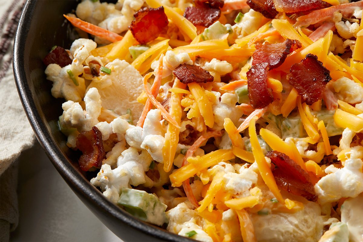 A close-up of a bowl filled with a salad made of cauliflower florets, shredded cheddar cheese, crispy bacon pieces, celery, and creamy dressing.