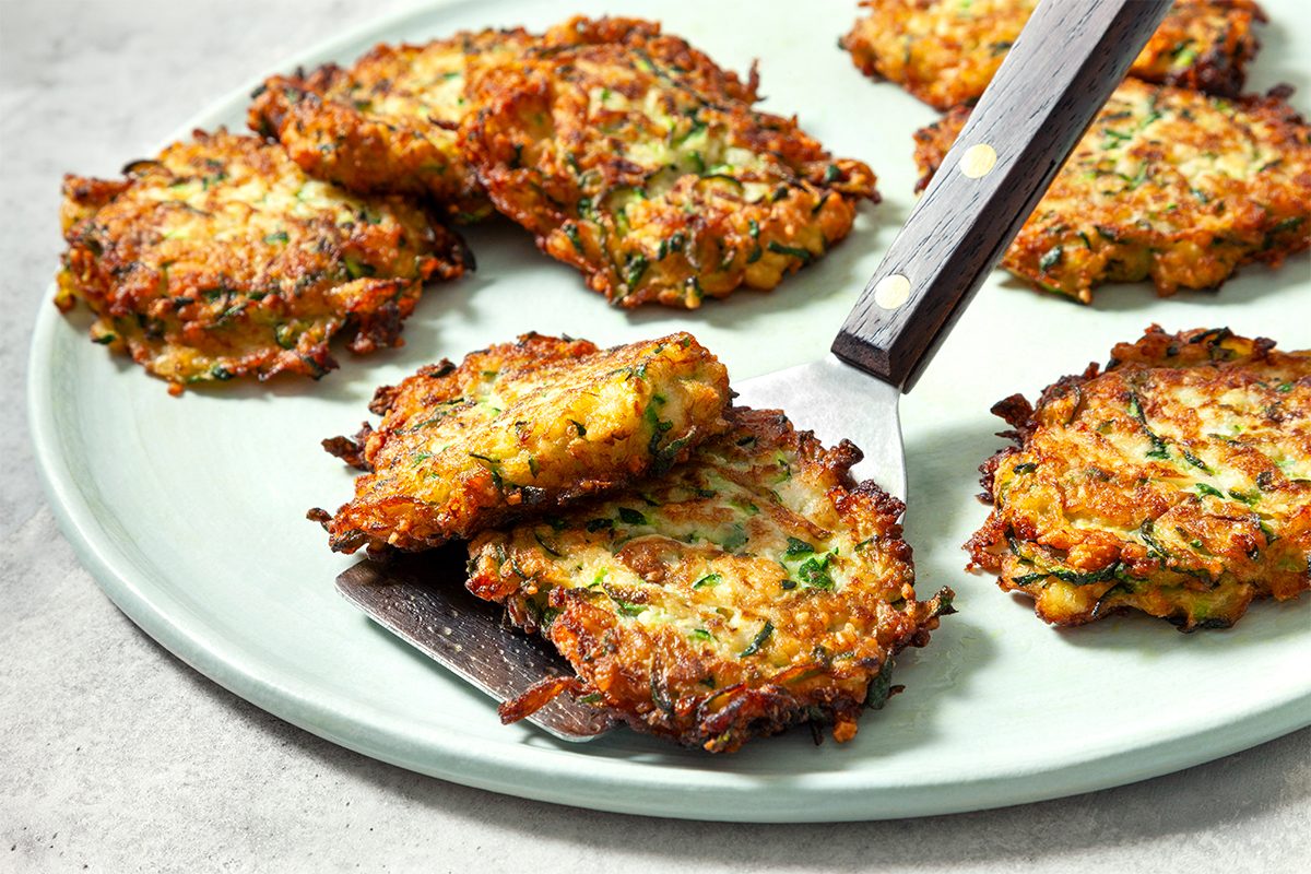 A plate of crispy, golden-brown zucchini fritters garnished with herbs. A spatula is lifting one fritter, showing the texture and green bits inside. The plate rests on a light-colored surface.